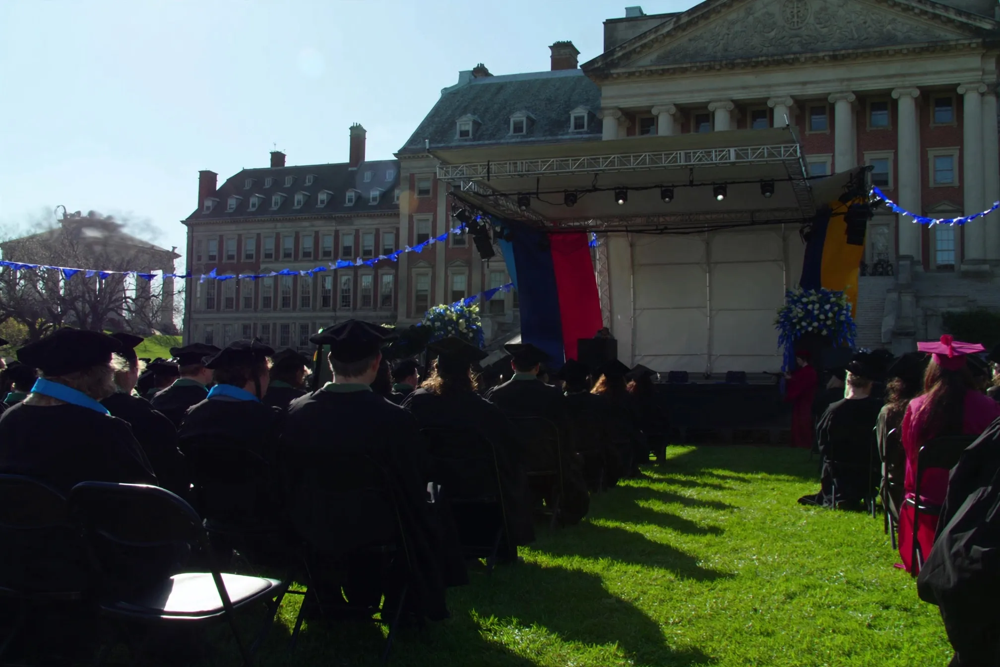A large convocation crowd of graduates in gowns seen from behind, seated on a green campus lawn facing a distant stage