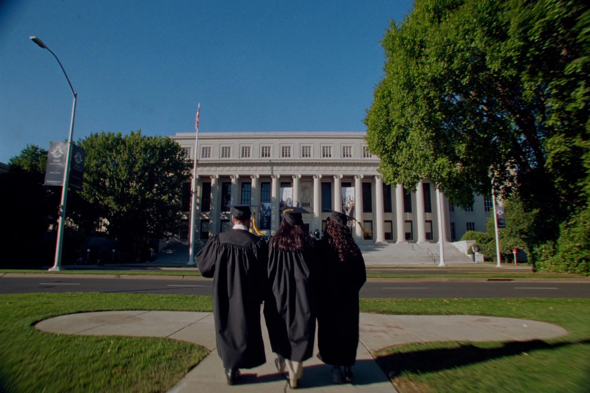 Three graduates in gowns and caps seen from behind, walking up a sunlit path toward a columned campus library