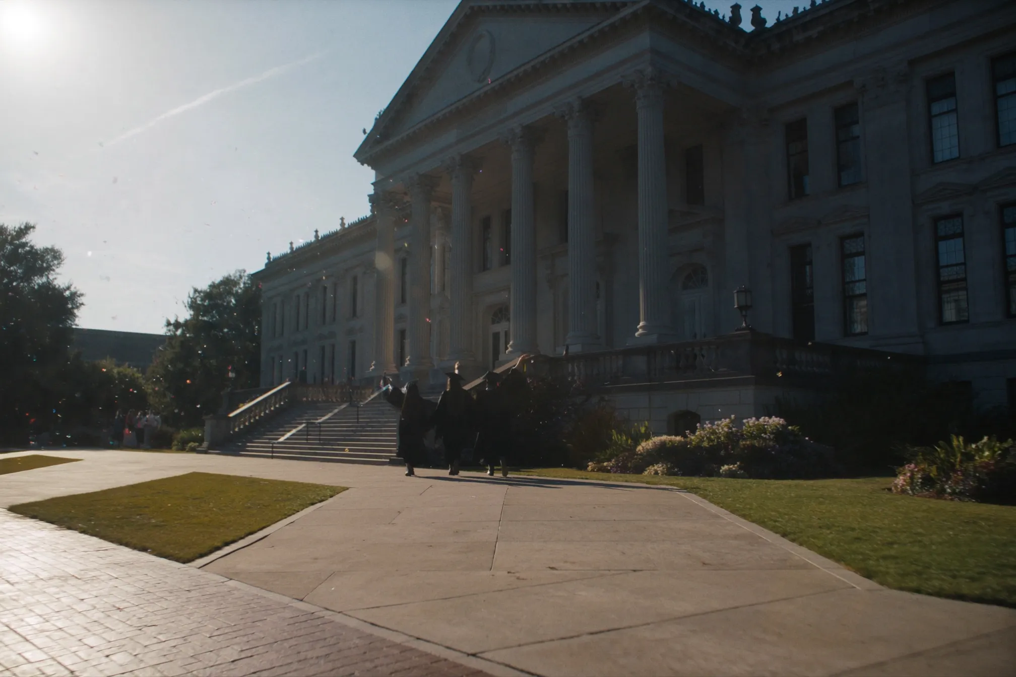 Three graduates in gowns seen from behind with arms raised, walking toward a columned university building